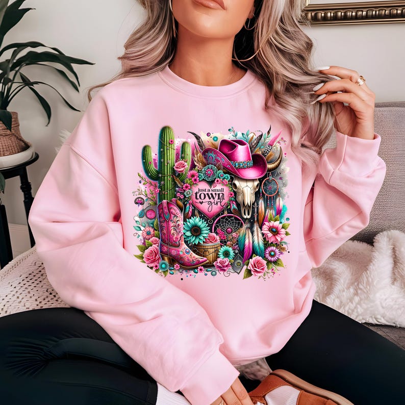 May include: Pink sweatshirt with a graphic design featuring a cactus, a cowboy hat, a skull, a boot, and flowers. The text "Just a small town girl" is printed on the graphic.