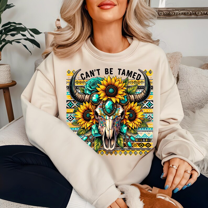 K&ouml;nnte beinhalten: Ein beiger Pullover mit einem Grafikdruck eines Sch&auml;dels mit H&ouml;rnern, umgeben von Sonnenblumen und t&uuml;rkisfarbenen Juwelen. Der Text "Can't Be Tamed" ist &uuml;ber dem Sch&auml;del gedruckt.