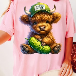 May include: Pink t-shirt with a cartoon Highland cow wearing a green baseball cap that says "Just Dill With It." The cow is holding a green pickle.