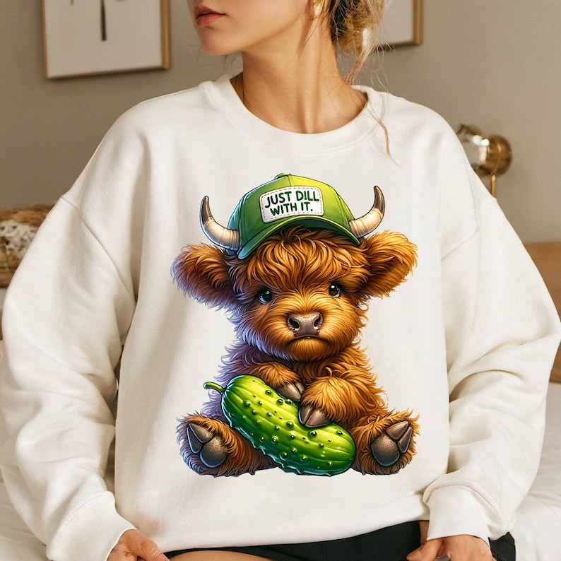May include: White sweatshirt with a cartoon illustration of a Highland cow wearing a green baseball cap that says "Just Dill With It." The cow is holding a green pickle.