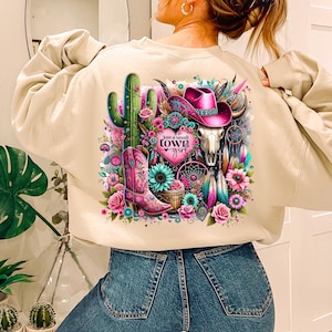 May include: Beige sweatshirt with a colorful graphic print featuring a cactus, a cowboy hat, a skull, and the text "Just a small town girl".