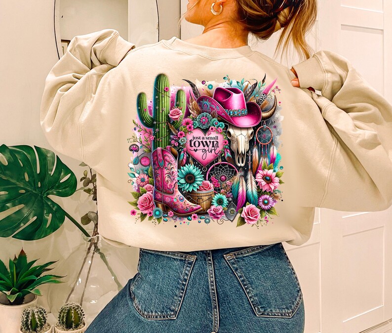 May include: Beige sweatshirt with a colorful graphic print featuring a cactus, a cowboy hat, a skull, and the text "Just a small town girl".