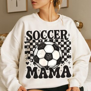 May include: Cream-colored sweatshirt with a black and white soccer ball graphic and the words "SOCCER MAMA" in a retro font. The design includes a checkered background, hearts, and star accents.