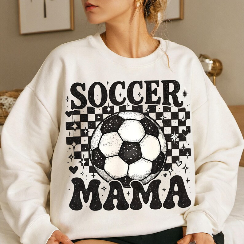 May include: Cream-colored sweatshirt with a black and white soccer ball graphic and the words "SOCCER MAMA" in a retro font. The design includes a checkered background, hearts, and star accents.
