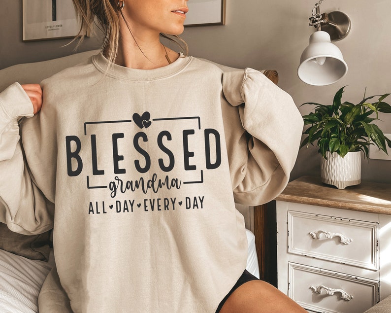 Op de afbeelding: Een beige sweatshirt met de tekst "Blessed grandma all day every day" in zwarte letters. Twee kleine hartjes bevinden zich boven het woord "Blessed".