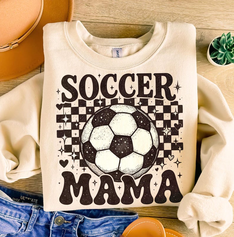 May include: Cream-colored sweatshirt with the words "SOCCER MAMA" in a retro font. A soccer ball graphic is centered on the shirt, set against a black and white checkered background with star and heart accents. The sweatshirt is folded.