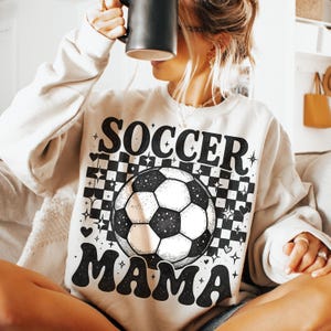 May include: Beige sweatshirt with the words "SOCCER MAMA" in black, featuring a soccer ball graphic. The design includes a black and white checkered pattern and star and heart accents. The sweatshirt has long sleeves.
