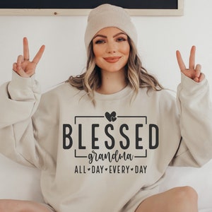 Op de afbeelding: Een beige sweatshirt met de tekst "BLESSED - grandma - ALL-DAY-EVERY-DAY" in zwarte letters. Twee kleine hartjes bevinden zich boven het woord "BLESSED".