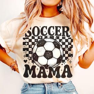 May include: Cream-colored t-shirt with the words "SOCCER MAMA" in black, retro-style lettering. A soccer ball graphic is centered above the word "MAMA". The background features a black and white checkered pattern with star and heart accents.