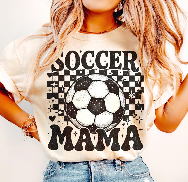 May include: Cream-colored t-shirt with the words "SOCCER MAMA" in black, retro-style lettering. A soccer ball graphic is centered above the word "MAMA". The background features a black and white checkered pattern with star and heart accents.
