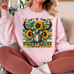 K&ouml;nnte beinhalten: Rosafarbener Pullover mit einem Aufdruck eines t&uuml;rkisfarbenen und goldenen Sch&auml;dels mit Sonnenblumen und dem Text "Can't Be Tamed".