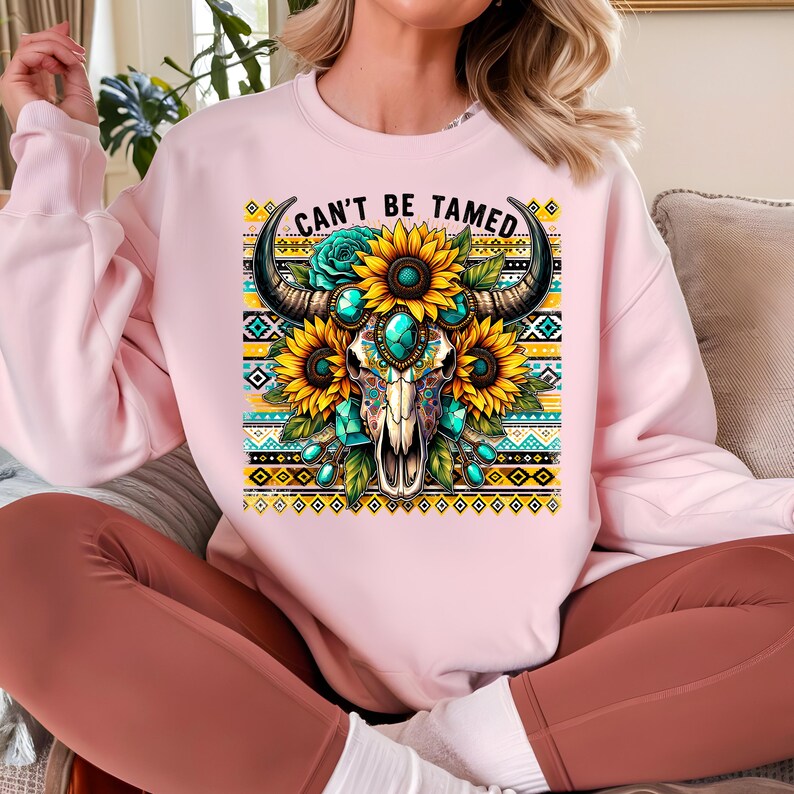K&ouml;nnte beinhalten: Rosafarbener Pullover mit einem Aufdruck eines t&uuml;rkisfarbenen und goldenen Sch&auml;dels mit Sonnenblumen und dem Text "Can't Be Tamed".