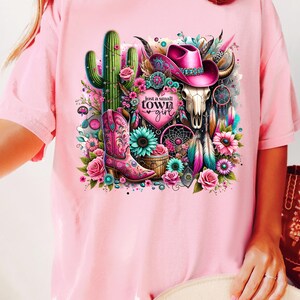 May include: Pink t-shirt with a graphic design of a cactus, a cowboy hat, a skull, a boot, and a dreamcatcher. The text "Just a small town girl" is on the graphic.