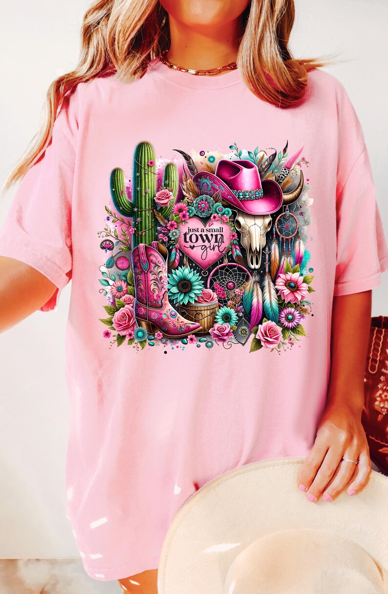 May include: Pink t-shirt with a graphic design of a cactus, a cowboy hat, a skull, a boot, and a dreamcatcher. The text "Just a small town girl" is on the graphic.