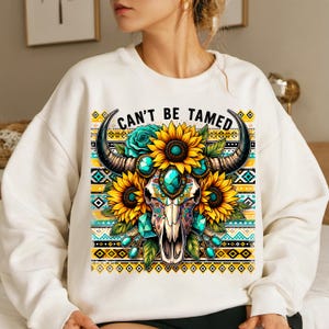 K&ouml;nnte beinhalten: Wei&szlig;er Pullover mit einem bunten Motiv eines Sch&auml;dels mit H&ouml;rnern, umgeben von Sonnenblumen und t&uuml;rkisfarbenen Edelsteinen. Der Text "Can't Be Tamed" ist &uuml;ber dem Motiv gedruckt.
