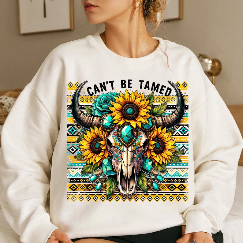 K&ouml;nnte beinhalten: Wei&szlig;er Pullover mit einem bunten Motiv eines Sch&auml;dels mit H&ouml;rnern, umgeben von Sonnenblumen und t&uuml;rkisfarbenen Edelsteinen. Der Text "Can't Be Tamed" ist &uuml;ber dem Motiv gedruckt.