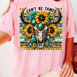 K&ouml;nnte beinhalten: Pinkes T-Shirt mit einem Aufdruck eines Sch&auml;dels mit t&uuml;rkisfarbenen Edelsteinen und Sonnenblumen. Der Sch&auml;del hat H&ouml;rner und ist von einem bunten geometrischen Muster umgeben. Der Text "Can't Be Tamed" ist auf das T-Shirt gedruckt.