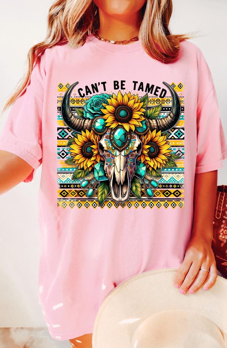 K&ouml;nnte beinhalten: Pinkes T-Shirt mit einem Aufdruck eines Sch&auml;dels mit t&uuml;rkisfarbenen Edelsteinen und Sonnenblumen. Der Sch&auml;del hat H&ouml;rner und ist von einem bunten geometrischen Muster umgeben. Der Text "Can't Be Tamed" ist auf das T-Shirt gedruckt.