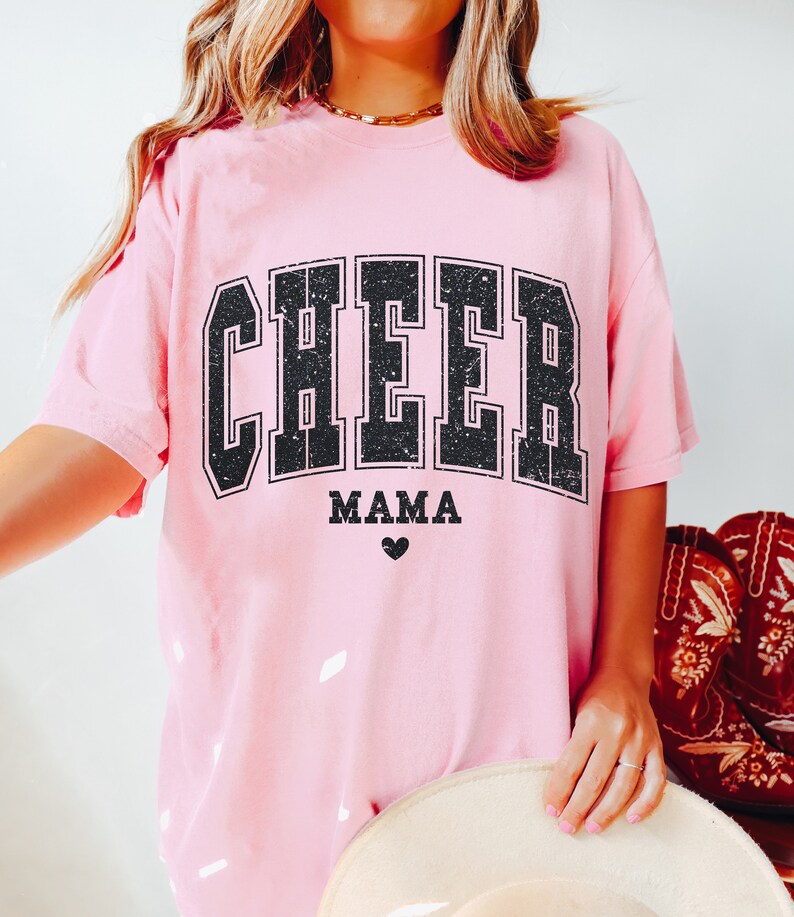 May include: Pink t-shirt with a black distressed graphic that says "CHEER MAMA" with a small heart below the text.