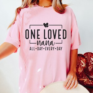 Może przedstawiać: R&oacute;żowa koszulka z czarnym nadrukiem, na kt&oacute;rym widnieje napis "One Loved Nana" z dwoma sercami nad tekstem i "All Day Every Day" pod tekstem.