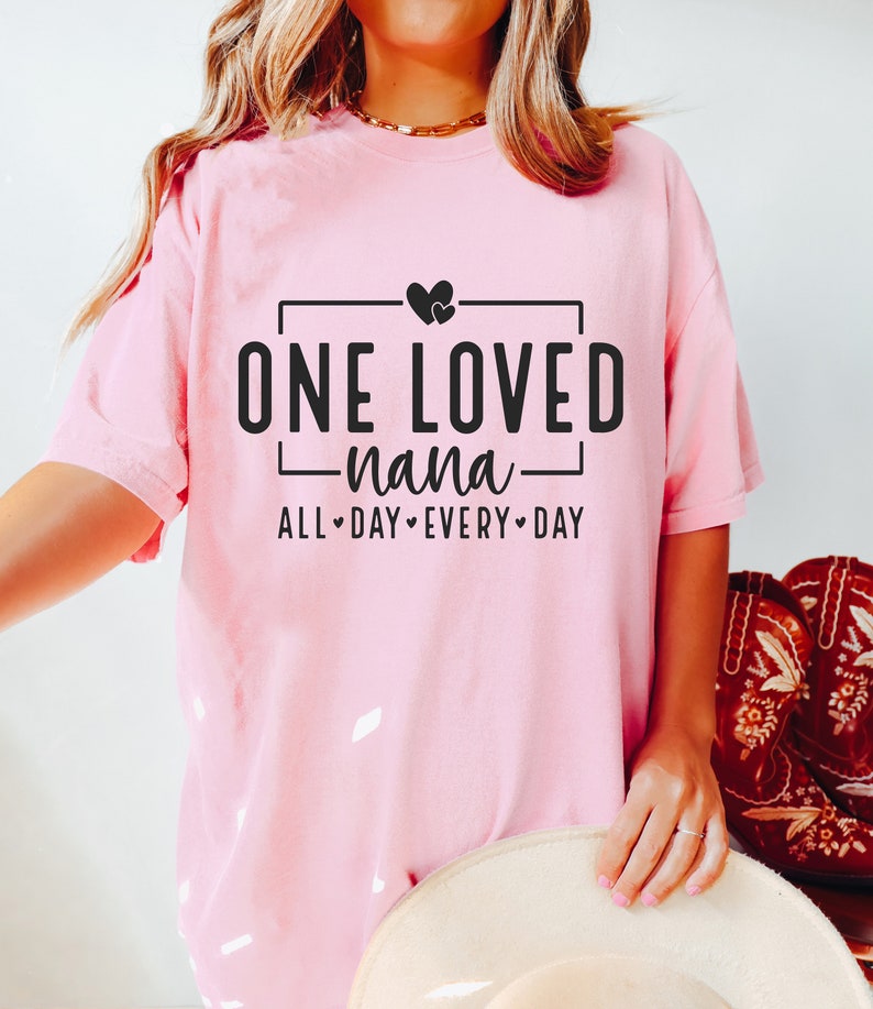 Może przedstawiać: R&oacute;żowa koszulka z czarnym nadrukiem, na kt&oacute;rym widnieje napis "One Loved Nana" z dwoma sercami nad tekstem i "All Day Every Day" pod tekstem.