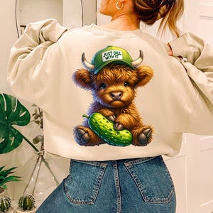 May include: A light beige sweatshirt with a cartoon illustration of a brown Highland cow holding a green dill pickle. The cow is wearing a green baseball cap that says "Just Dill With It."
