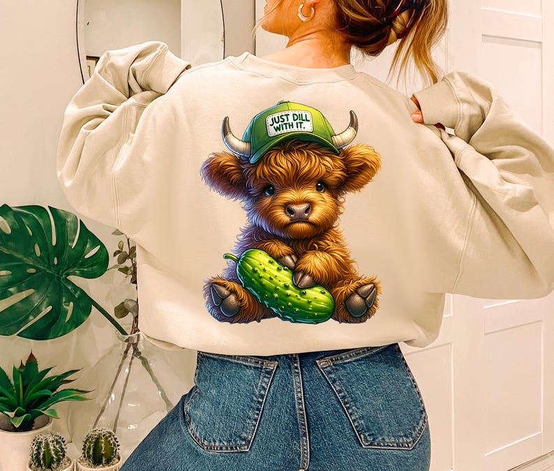 May include: A light beige sweatshirt with a cartoon illustration of a brown Highland cow holding a green dill pickle. The cow is wearing a green baseball cap that says "Just Dill With It."