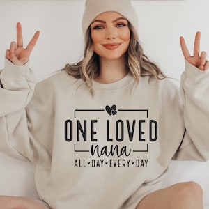 Może przedstawiać: Beżowa bluza z napisem "ONE LOVED nana ALL DAY EVERY DAY" w czarnym kolorze. Tekst jest umieszczony w czarnym prostokącie i zawiera dwa małe serduszka nad tekstem.