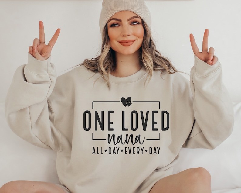 Może przedstawiać: Beżowa bluza z napisem "ONE LOVED nana ALL DAY EVERY DAY" w czarnym kolorze. Tekst jest umieszczony w czarnym prostokącie i zawiera dwa małe serduszka nad tekstem.