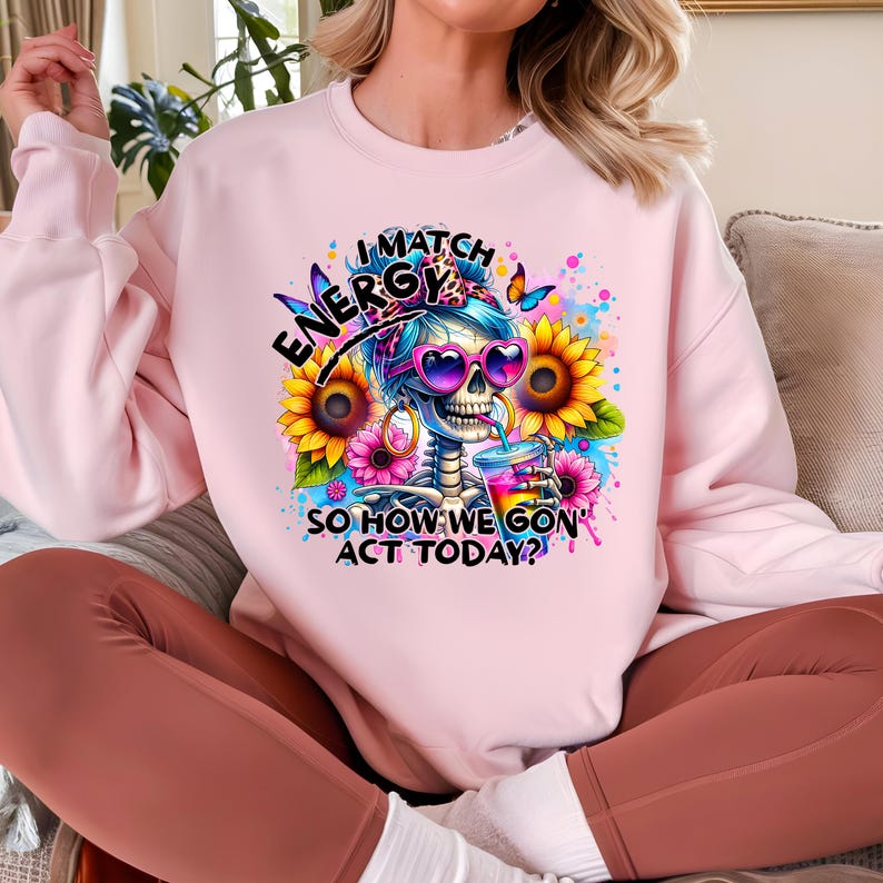 May include: Pink sweatshirt with a colorful graphic of a skeleton wearing sunglasses and holding a drink. The graphic says "I Match Energy So How We Gon' Act Today?"