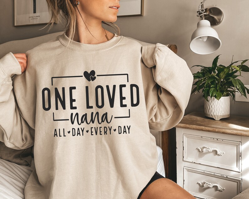 Może przedstawiać: Beżowa bluza z napisem "ONE LOVED -nana- ALL DAY EVERY DAY" w czarnym kolorze. Nad tekstem znajdują się dwa małe serduszka.