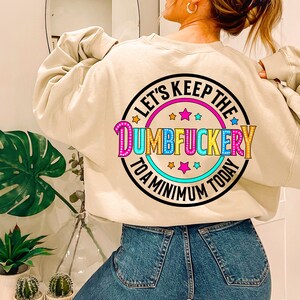 May include: A beige sweatshirt with a colorful graphic print that says "Let's Keep the Dumbfuckery to a Minimum Today".