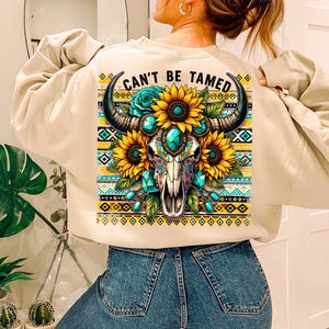K&ouml;nnte beinhalten: Ein beiger Pullover mit einem bunten Grafikdruck eines Sch&auml;dels mit t&uuml;rkisfarbenen Juwelen und Sonnenblumen. Der Text "Can't Be Tamed" ist auf dem Pullover aufgedruckt.