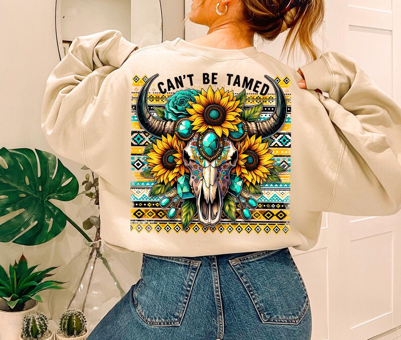 K&ouml;nnte beinhalten: Ein beiger Pullover mit einem bunten Grafikdruck eines Sch&auml;dels mit t&uuml;rkisfarbenen Juwelen und Sonnenblumen. Der Text "Can't Be Tamed" ist auf dem Pullover aufgedruckt.
