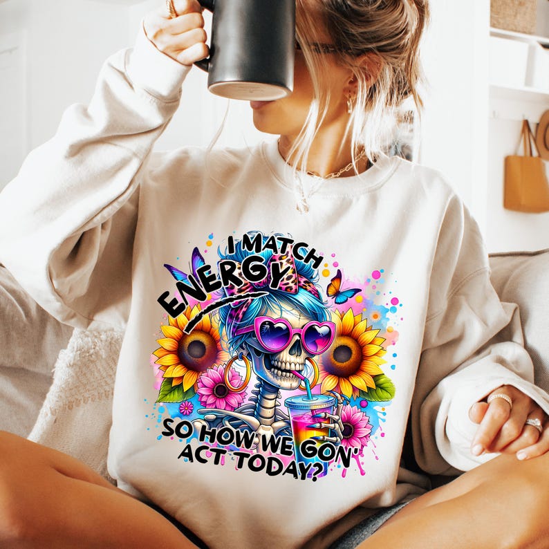 May include: White sweatshirt with a colorful graphic of a skeleton wearing sunglasses and a bow. The text on the sweatshirt reads "I Match Energy So How We Gon' Act Today?"
