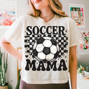 May include: Cream-colored t-shirt with the words "SOCCER MAMA" in a retro font. A black and white soccer ball is centered above the word "MAMA". The background features a checkerboard pattern with stars and hearts.