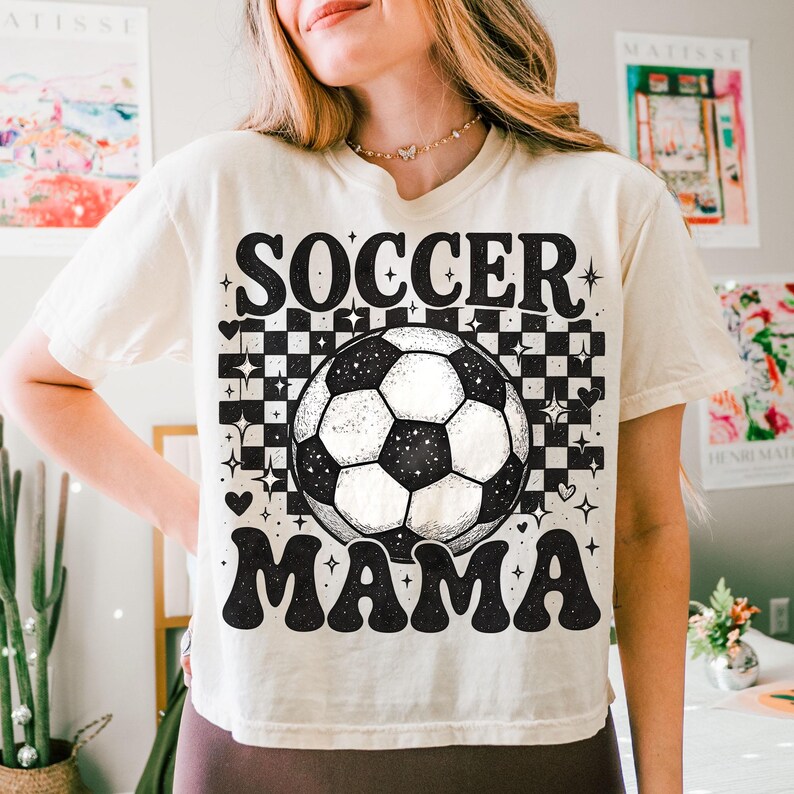 May include: Cream-colored t-shirt with the words "SOCCER MAMA" in a retro font. A black and white soccer ball is centered above the word "MAMA". The background features a checkerboard pattern with stars and hearts.