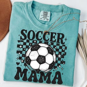 May include: A turquoise Comfort Colors t-shirt with the words "SOCCER MAMA" in black, with a soccer ball graphic. The shirt has a checkerboard pattern behind the soccer ball, with stars and hearts. A gold necklace is draped across the shirt.