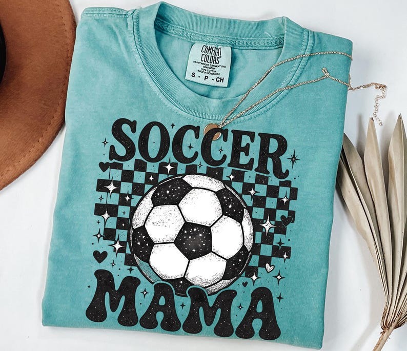 May include: A turquoise Comfort Colors t-shirt with the words "SOCCER MAMA" in black, with a soccer ball graphic. The shirt has a checkerboard pattern behind the soccer ball, with stars and hearts. A gold necklace is draped across the shirt.