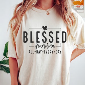Op de afbeelding: Een wit T-shirt met de tekst "Blessed grandma all day every day" in zwarte letters. Twee kleine hartjes bevinden zich boven het woord "Blessed".