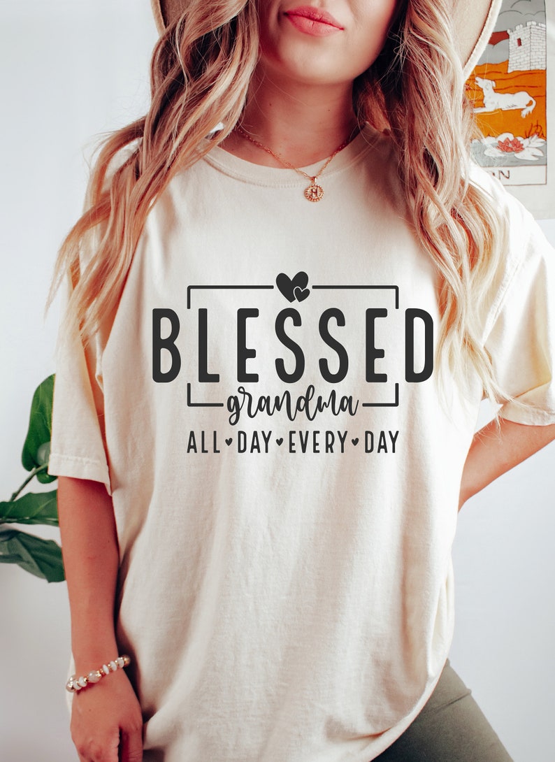 Op de afbeelding: Een wit T-shirt met de tekst "Blessed grandma all day every day" in zwarte letters. Twee kleine hartjes bevinden zich boven het woord "Blessed".