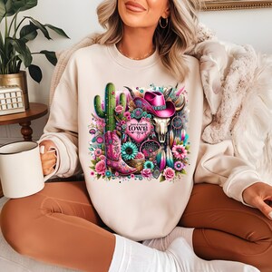 May include: A cream-colored sweatshirt with a graphic print of a cactus, a cowboy hat, a skull, a dreamcatcher, and a pair of cowboy boots. The graphic is surrounded by pink, blue, and green flowers. The text "Just a small town girl" is printed on the graphic.