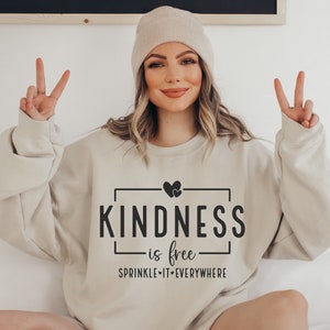 May include: A beige sweatshirt with the text "KINDNESS is free" and "SPRINKLE IT EVERYWHERE" in black. The sweatshirt features a small heart graphic above the text.