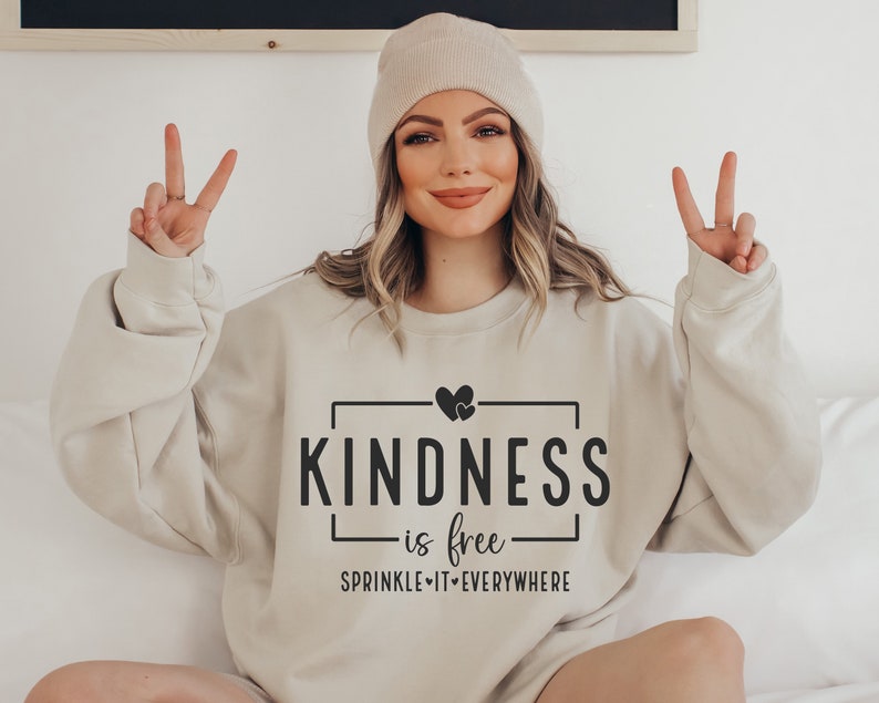 May include: A beige sweatshirt with the text "KINDNESS is free" and "SPRINKLE IT EVERYWHERE" in black. The sweatshirt features a small heart graphic above the text.