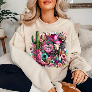 May include: A beige sweatshirt with a graphic print of a cactus, a pink cowboy hat, a skull, and a dreamcatcher. The graphic also includes the text "Just a small town girl".