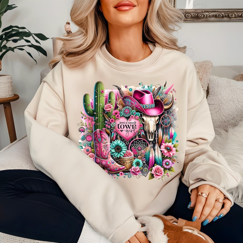 May include: A beige sweatshirt with a graphic print of a cactus, a pink cowboy hat, a skull, and a dreamcatcher. The graphic also includes the text "Just a small town girl".