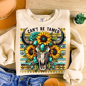 K&ouml;nnte beinhalten: Ein cremefarbener Pullover mit einem Aufdruck eines Sch&auml;dels mit H&ouml;rnern, umgeben von Sonnenblumen und t&uuml;rkisfarbenen Edelsteinen. Der Text "Can't Be Tamed" ist &uuml;ber dem Aufdruck gedruckt.