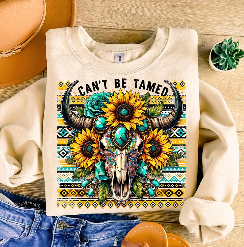 K&ouml;nnte beinhalten: Ein cremefarbener Pullover mit einem Aufdruck eines Sch&auml;dels mit H&ouml;rnern, umgeben von Sonnenblumen und t&uuml;rkisfarbenen Edelsteinen. Der Text "Can't Be Tamed" ist &uuml;ber dem Aufdruck gedruckt.