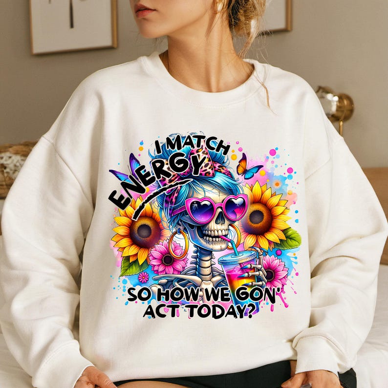 May include: White sweatshirt with a colorful graphic of a skeleton with a flower crown and sunglasses. The text reads "I match energy so how we gon' act today?"