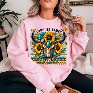 K&ouml;nnte beinhalten: Rosafarbener Pullover mit einem bunten Grafikdruck eines Stierssch&auml;dels mit Sonnenblumen und t&uuml;rkisfarbenen Juwelen. Der Text "Can't Be Tamed" ist &uuml;ber dem Grafikdruck gedruckt.